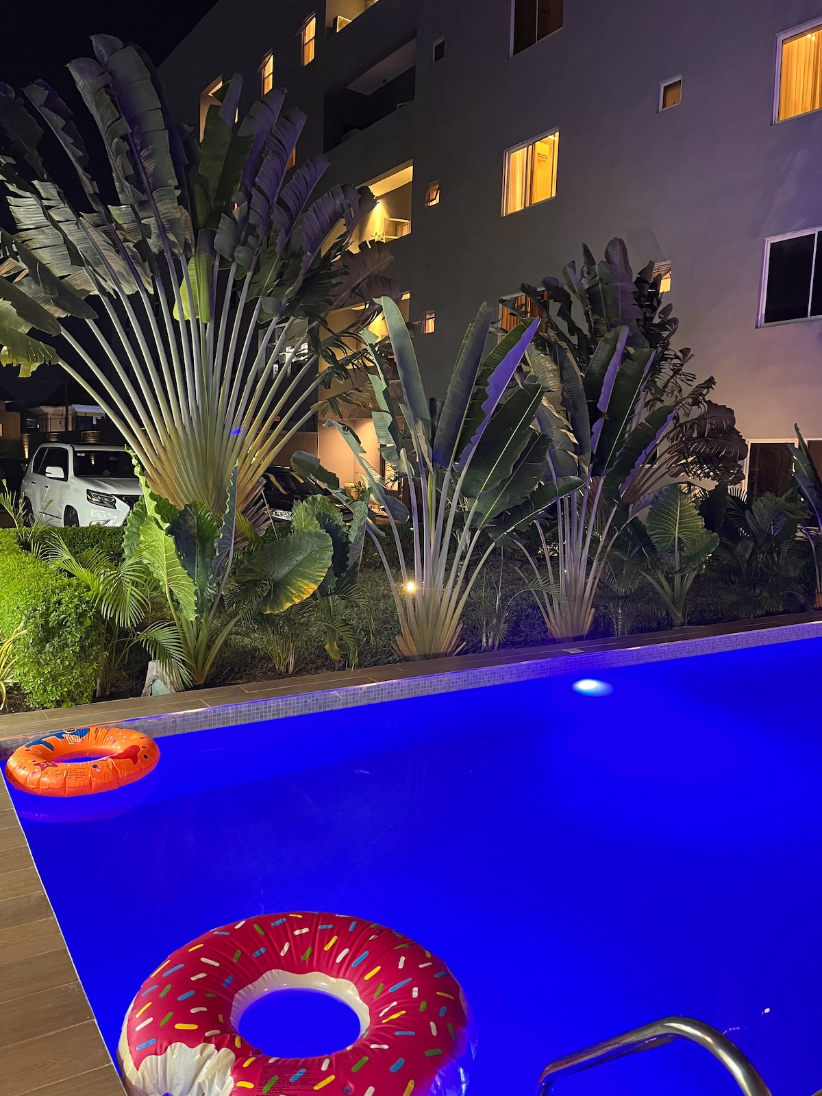 The image captures a swimming pool with illuminated blue water, surrounded by lush greenery. Inflatable rings in bright colors float on the surface. Light from nearby windows reflects off the water, enhancing the tranquil evening atmosphere of the landscaped area.