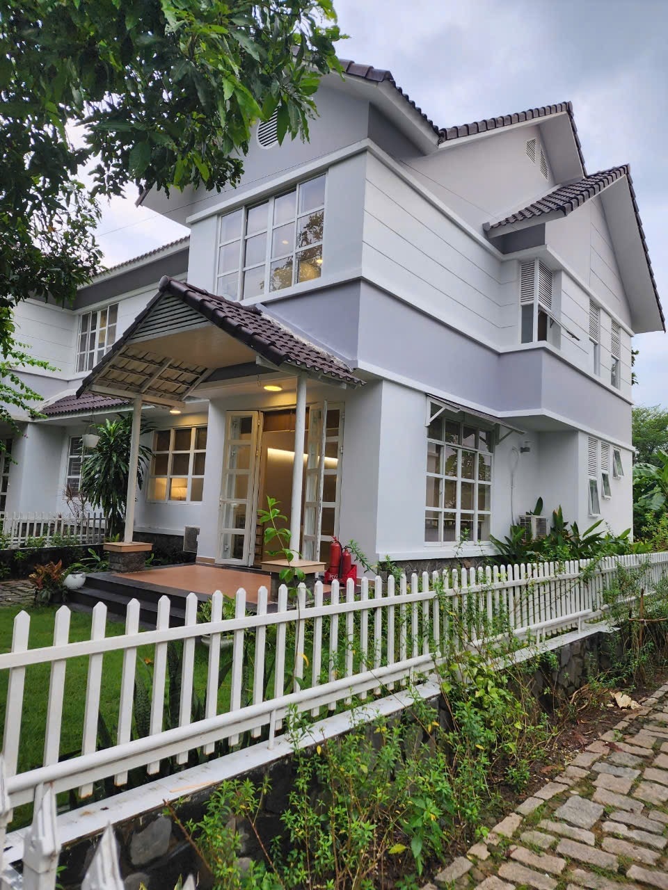 A two-story house is shown, featuring a light-colored exterior with a contrast of dark roofing. Large windows allow natural light to fill the interior. A small porch area is visible, adorned with plants and a white picket fence that encloses the garden.