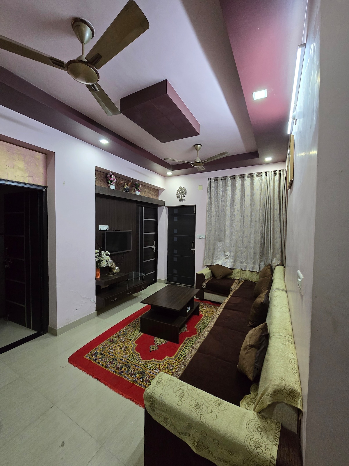 A cozy living area is presented with a large brown sectional sofa and a low coffee table positioned on a decorative rug. Wall-mounted shelves offer a space for decor, while the room is brightened by overhead ceiling fans and natural light filtering through the window behind elegant curtains.