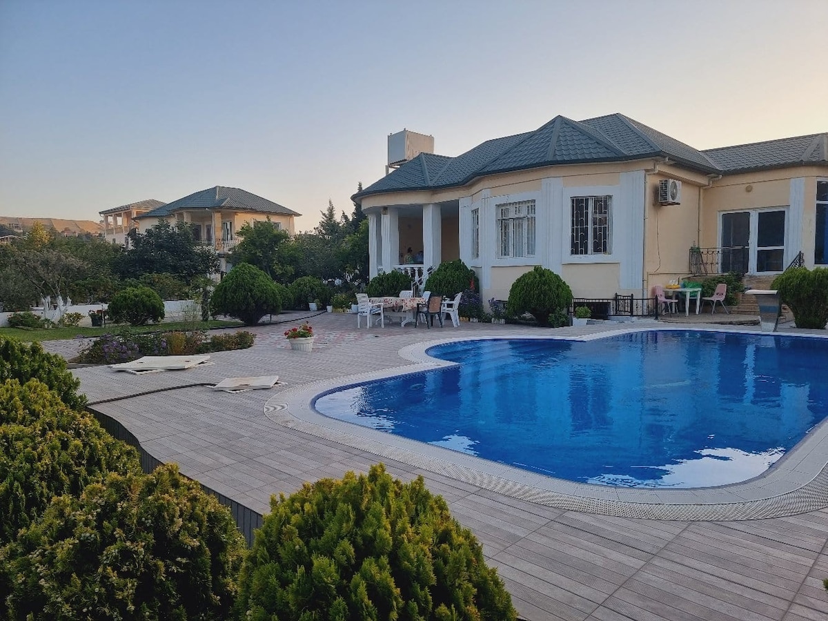 An outdoor pool is framed by neatly landscaped greenery and wooden decking. The house features a spacious terrace with seating areas, inviting relaxation. Lush bushes create natural boundaries, enhancing the tranquil environment, while the sky transitions to evening hues, providing a serene backdrop.