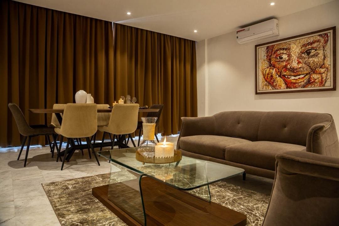 A modern living area is presented, featuring a plush sofa and a glass coffee table atop a soft rug. An elegant dining table is set for four, surrounded by comfortable chairs. Warm lighting and rich brown curtains create a cozy ambiance.
