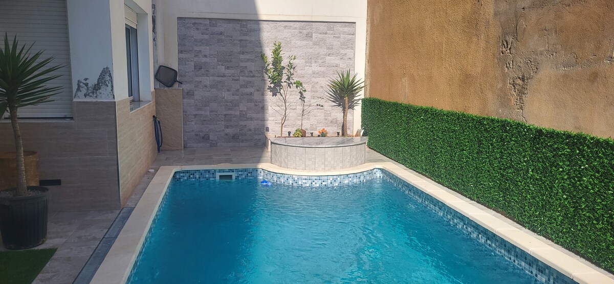 A private terrace features a rectangular swimming pool, surrounded by tall greenery and potted plants. The pool's clear blue water reflects sunlight, enhancing the serene atmosphere. A textured wall serves as a backdrop, contrasting with the smooth pool tiles and greenery.