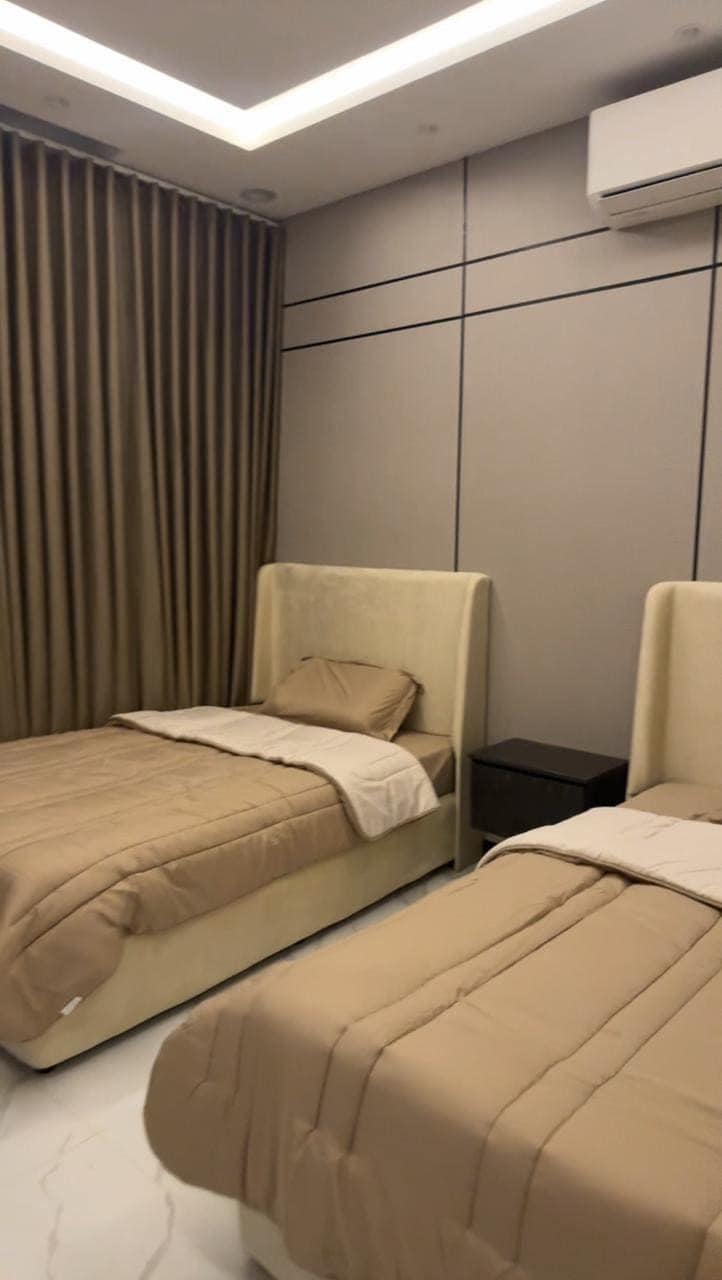 The sleeping area features two light-toned beds with headboards, dressed in neutral bedding. A nightstand is positioned between the beds, and textured curtains cover a wall, contributing to the modern aesthetic of the room.