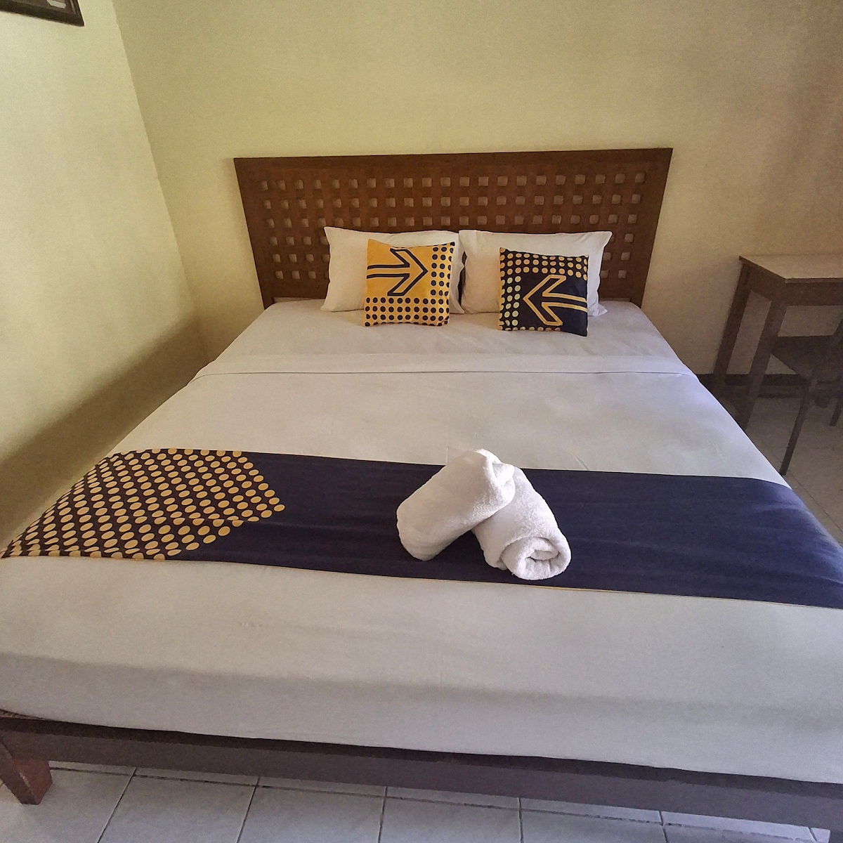 A spacious bed is presented with patterned decorative pillows and a contrasting blanket. Fresh towels are neatly arranged on the bed. A simple wooden table is positioned nearby, contributing to the clean and minimal aesthetic of the room.
