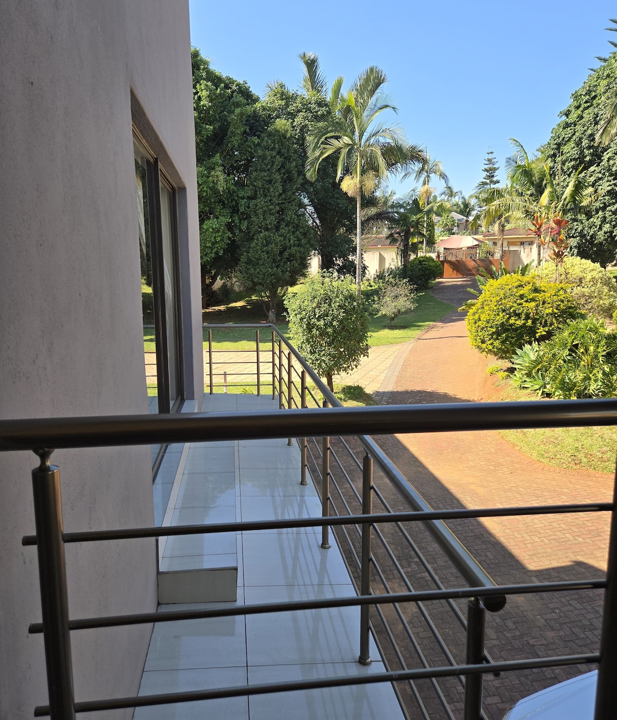 A view from the balcony showcases a well-maintained garden with lush greenery and palm trees. A winding pathway is visible, leading through the landscape, while a clear blue sky completes the serene outdoor scene.