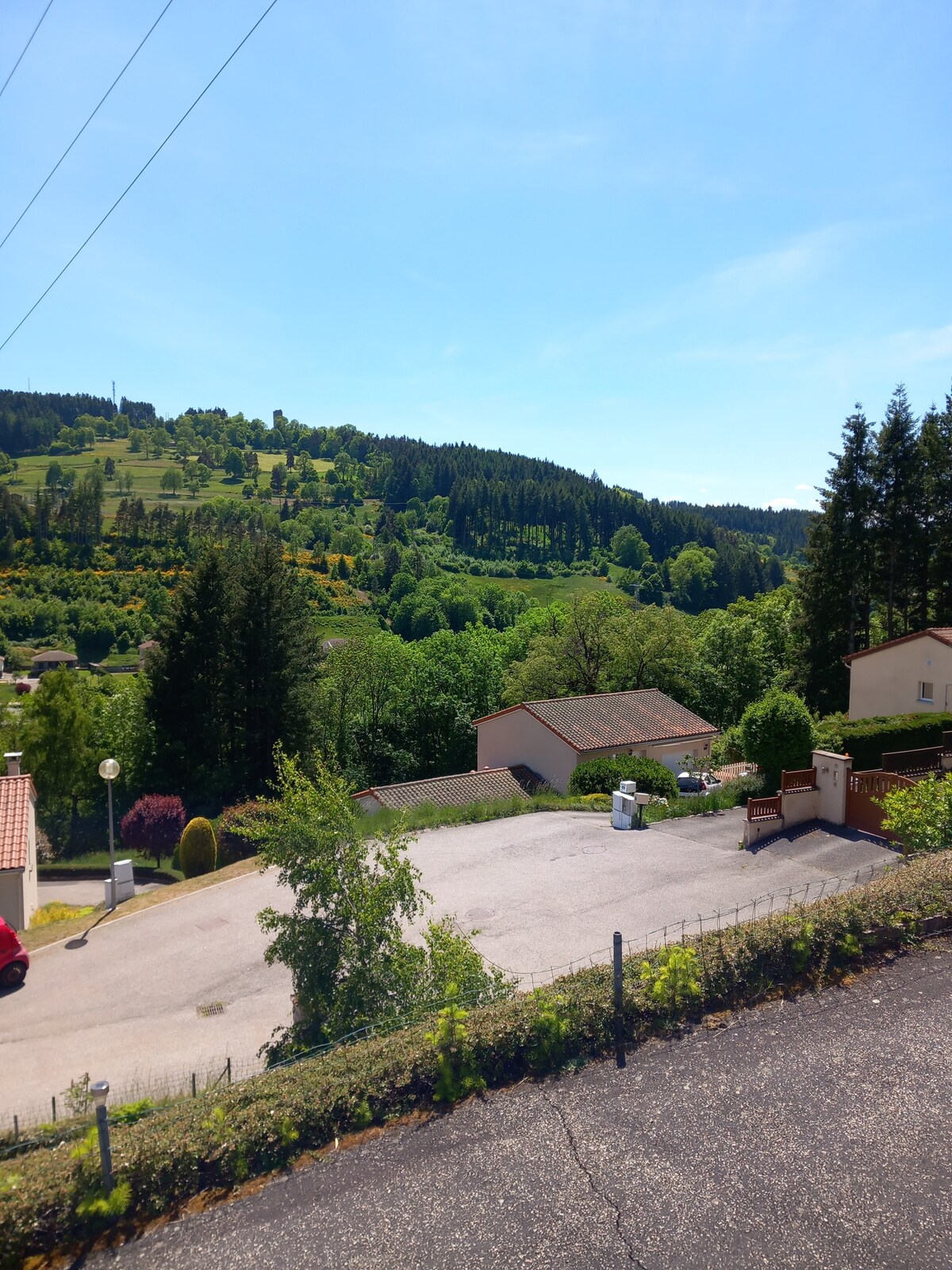 A serene landscape is depicted, showcasing rolling green hills and dense trees under a clear blue sky. A mix of residential buildings and greenery is visible, along with a well-maintained road that winds through the area, contributing to a sense of tranquility.