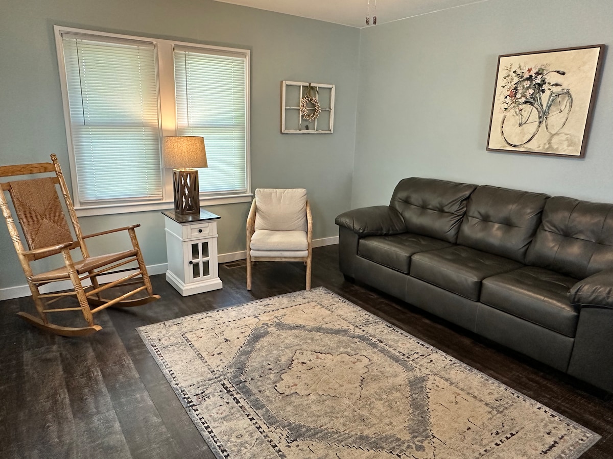A welcoming living area features a comfortable leather sofa and a light-colored armchair. A wooden rocking chair is positioned by a bright window with soft, natural light filtering through. A decorative area rug covers the dark floor, while a small lamp and wall art add subtle details.