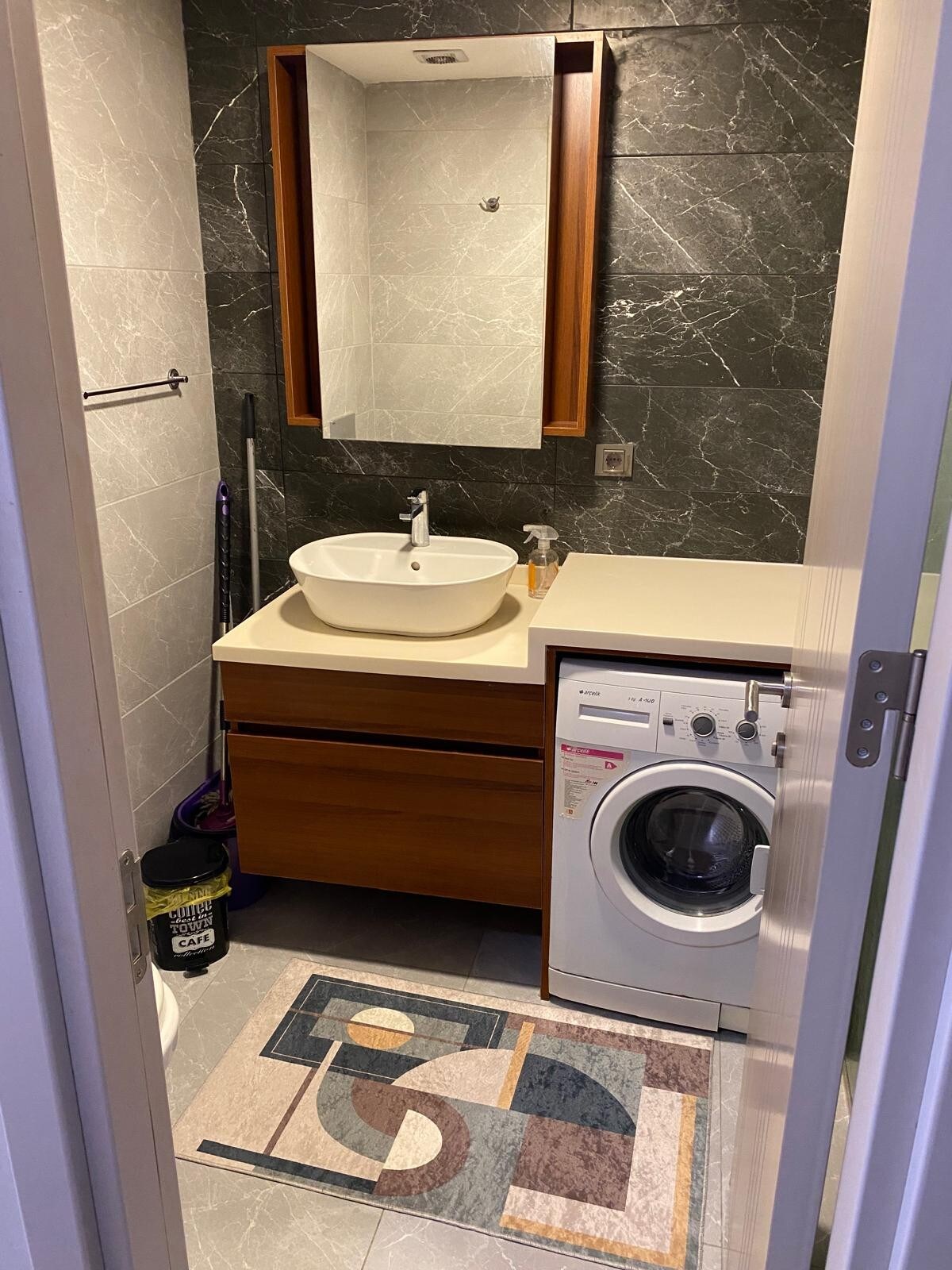 A compact bathroom featuring modern fixtures and a stylish design. A vessel sink is positioned on a wooden vanity, complemented by a mirrored cabinet. A washing machine is included, and a patterned rug is laid on the tiled floor, creating a functional and organized space.