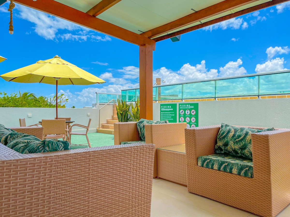 A shared leisure area is displayed, featuring woven seating with tropical-patterned cushions. Two large yellow umbrellas provide shade over a table and chairs. A staircase leads to the pool area, accompanied by green plants and a backdrop of blue skies with clouds.