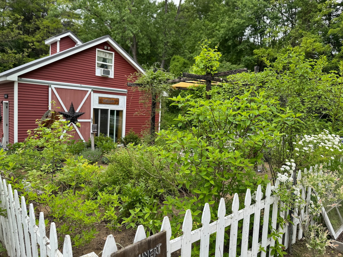 A charming red barn is surrounded by lush greenery and flowering plants. A white picket fence encloses the outdoor area, with a small garden pathway visible. Above, a shaded seating area is indicated by an outdoor structure, inviting guests to enjoy nature.
