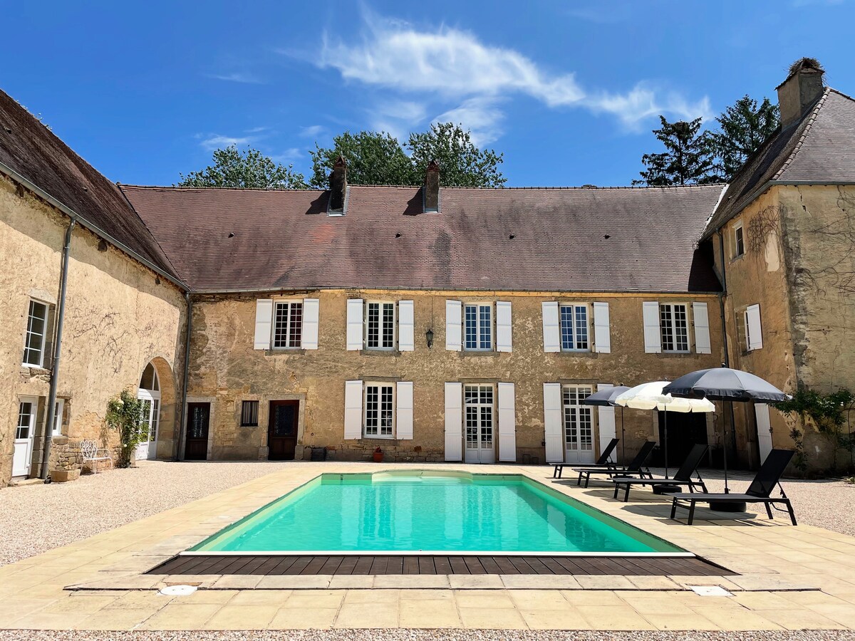 A spacious outdoor area is highlighted by a rectangular swimming pool surrounded by a light-colored patio. Several lounge chairs with umbrellas provide shaded seating. The building's rustic stone façade and white shuttered windows create a harmonious backdrop against the clear blue sky.