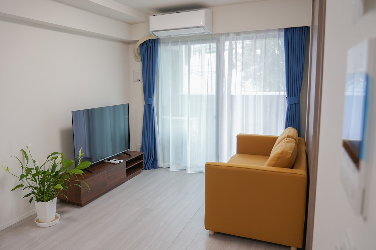 A bright living area features a large window adorned with sheer curtains offering natural light. A yellow couch is positioned opposite a wall-mounted television, and a potted plant adds a touch of greenery. Light wood flooring creates a clean, spacious feel.