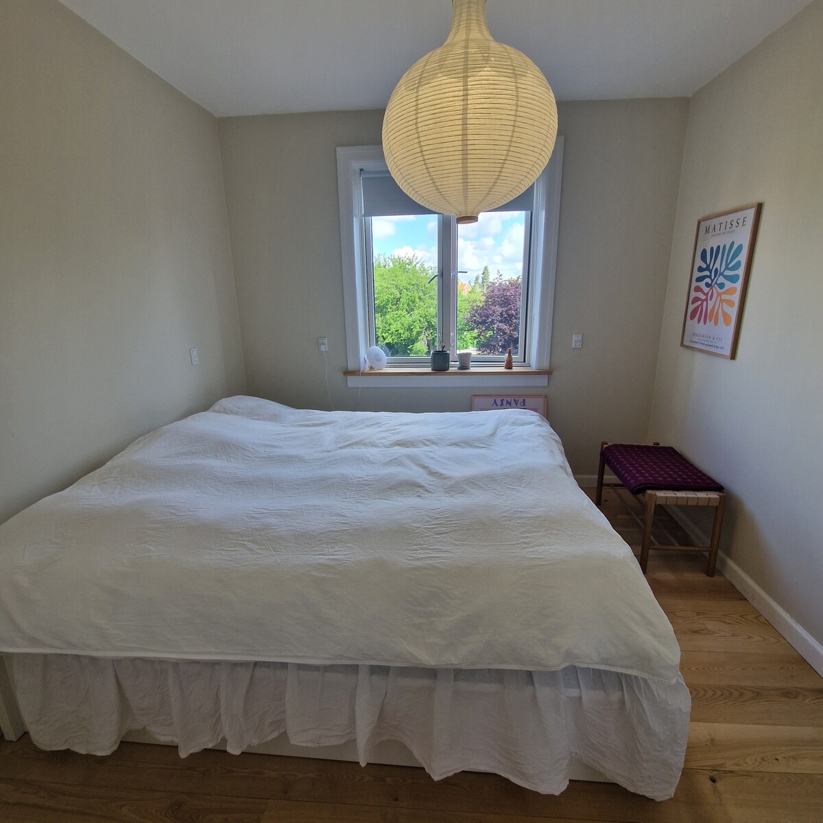 A spacious bedroom features a large, neatly made bed with a white duvet cover. A round pendant light hangs above, providing soft illumination. A window offers a view of greenery outside, while a small, decorative chair is positioned nearby.