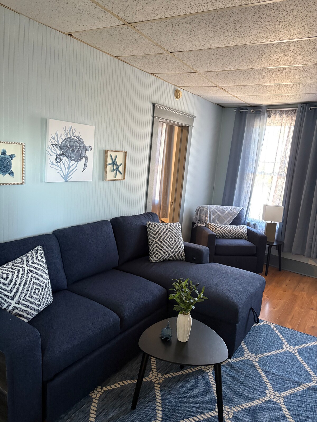 A comfortable seating area is showcased, featuring a dark blue sectional couch adorned with patterned throw pillows. A small black table sits in front, complemented by a decorative plant. Natural light enters through a window, with soft gray curtains framing the view.