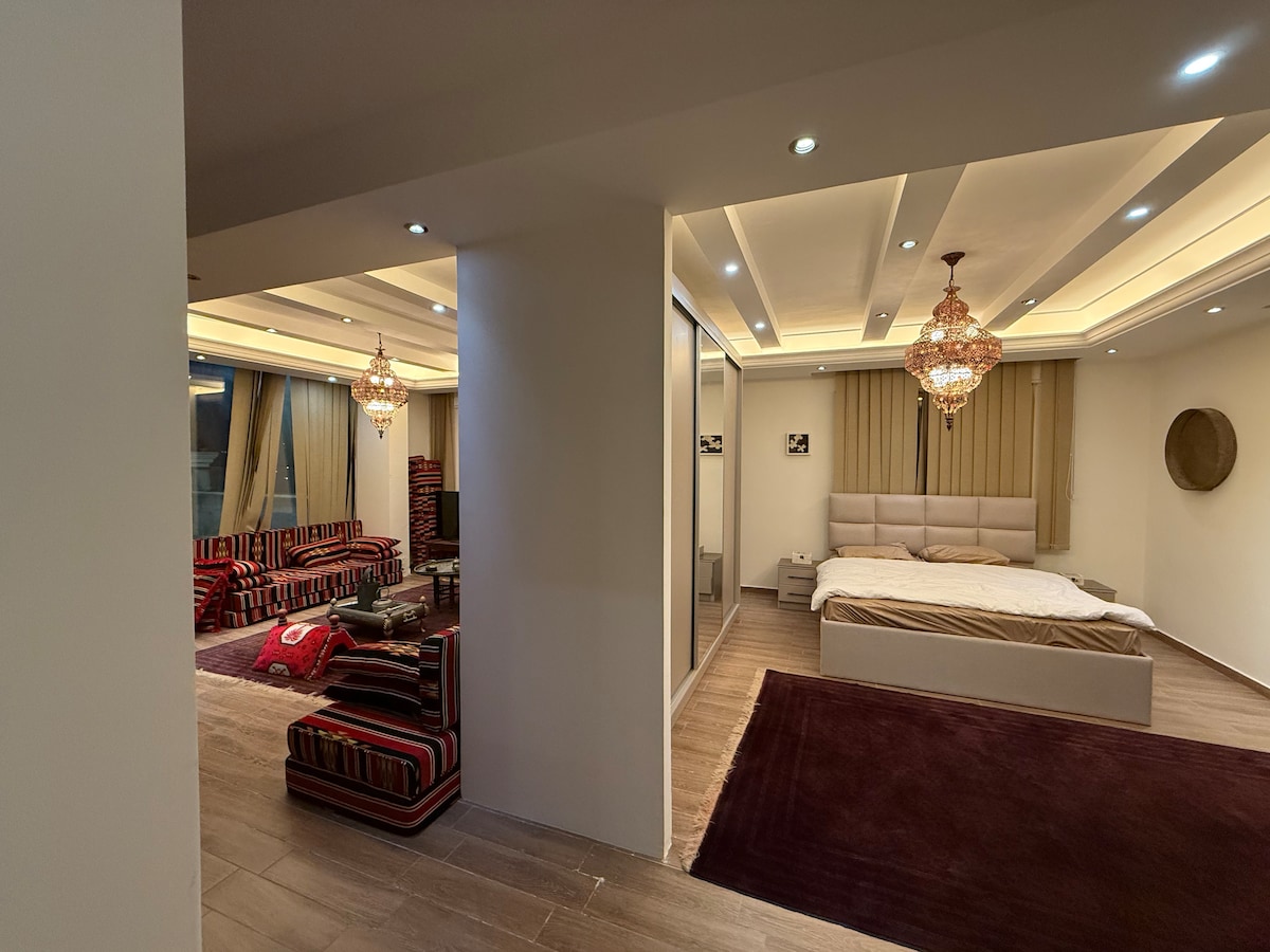 A spacious living area is visible, adorned with traditional seating and warm lighting from ornate chandeliers. A comfortable bed is seen in an adjoining room, complemented by soft bedding. Natural light filters through large windows, enhancing the elegant design of the space.