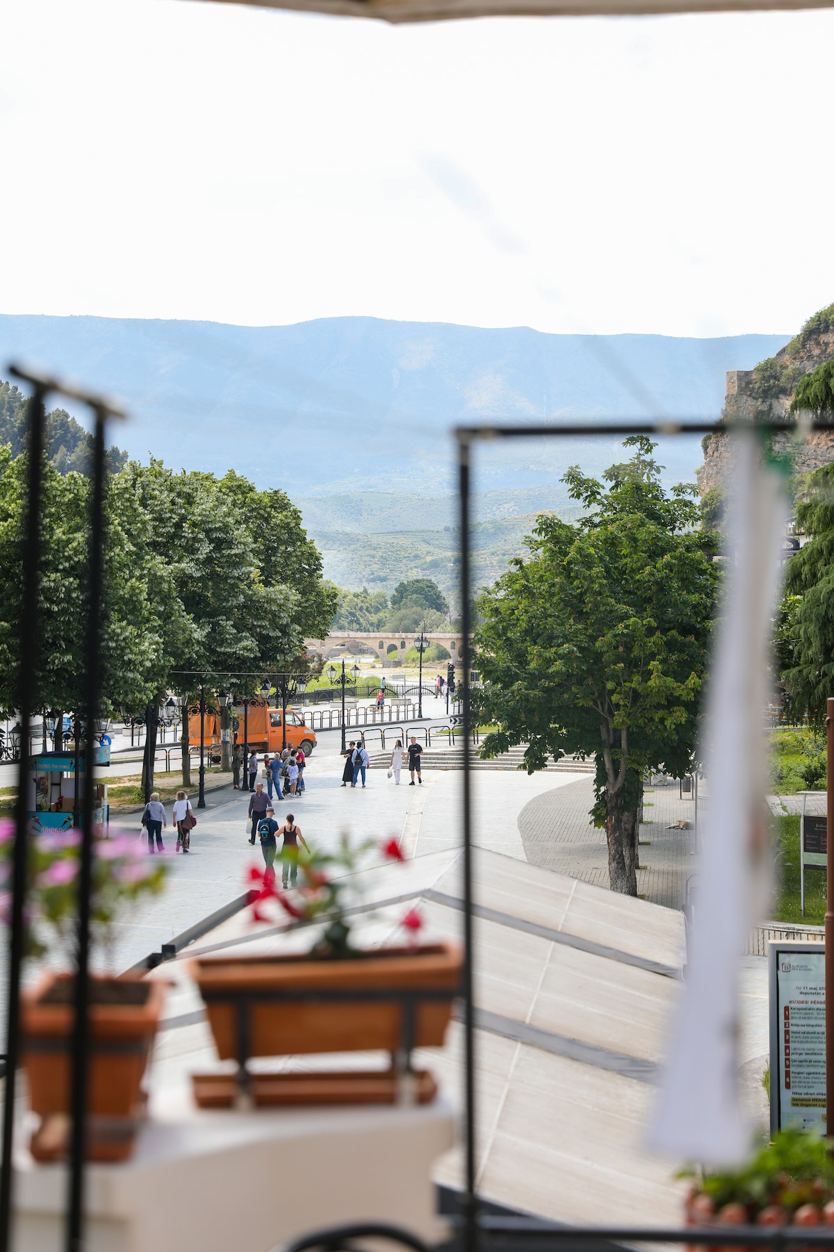 A view is captured from a balcony, featuring a wide street lined with trees and colorful flowers in pots. People are seen walking along the pathway, with mountains visible in the background under a partly cloudy sky.