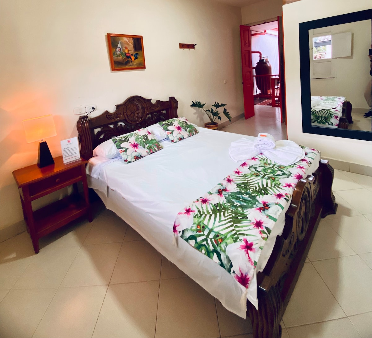 A spacious bed is adorned with floral-patterned linens, complemented by decorative pillows. A wooden nightstand with a lamp is positioned beside the bed. A large mirror reflects the room, which features a minimalistic design and neutral-colored walls.