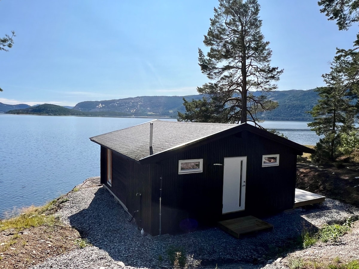 A modern cabin with a dark exterior is positioned near the tranquil waters of Lake Nisser. The landscape features open skies and surrounding trees, providing a sense of privacy and seclusion. A small outdoor platform extends from the cabin, enhancing the outdoor living space.