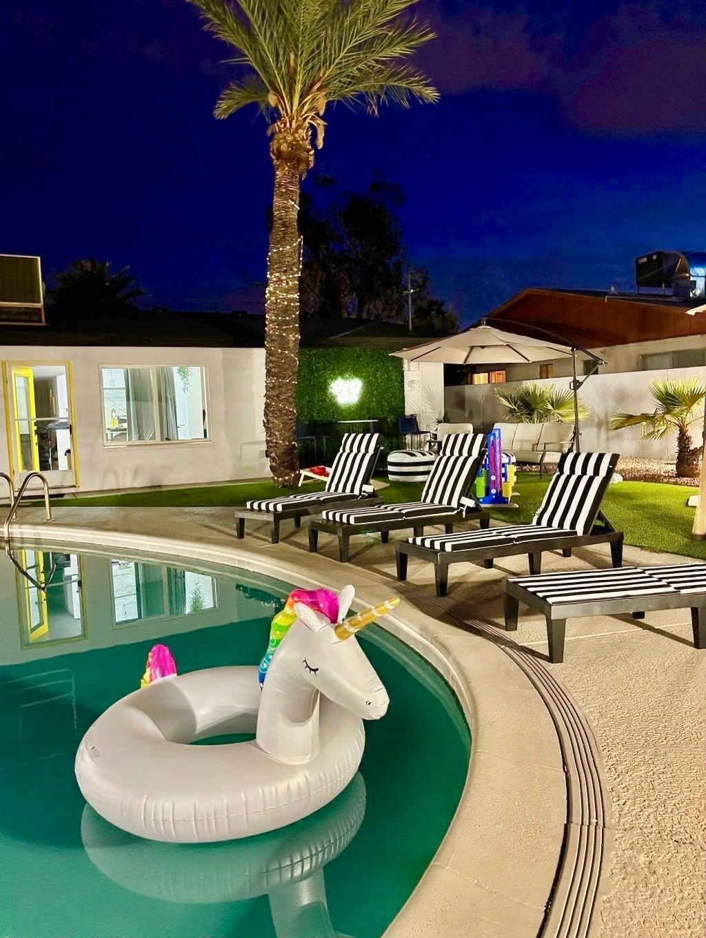 A well-lit backyard scene features a large, inviting pool surrounded by striped loungers. A whimsical inflatable unicorn floats in the water, while palm trees provide a tropical ambiance. Soft lighting enhances the comfortable atmosphere, perfect for evening relaxation.