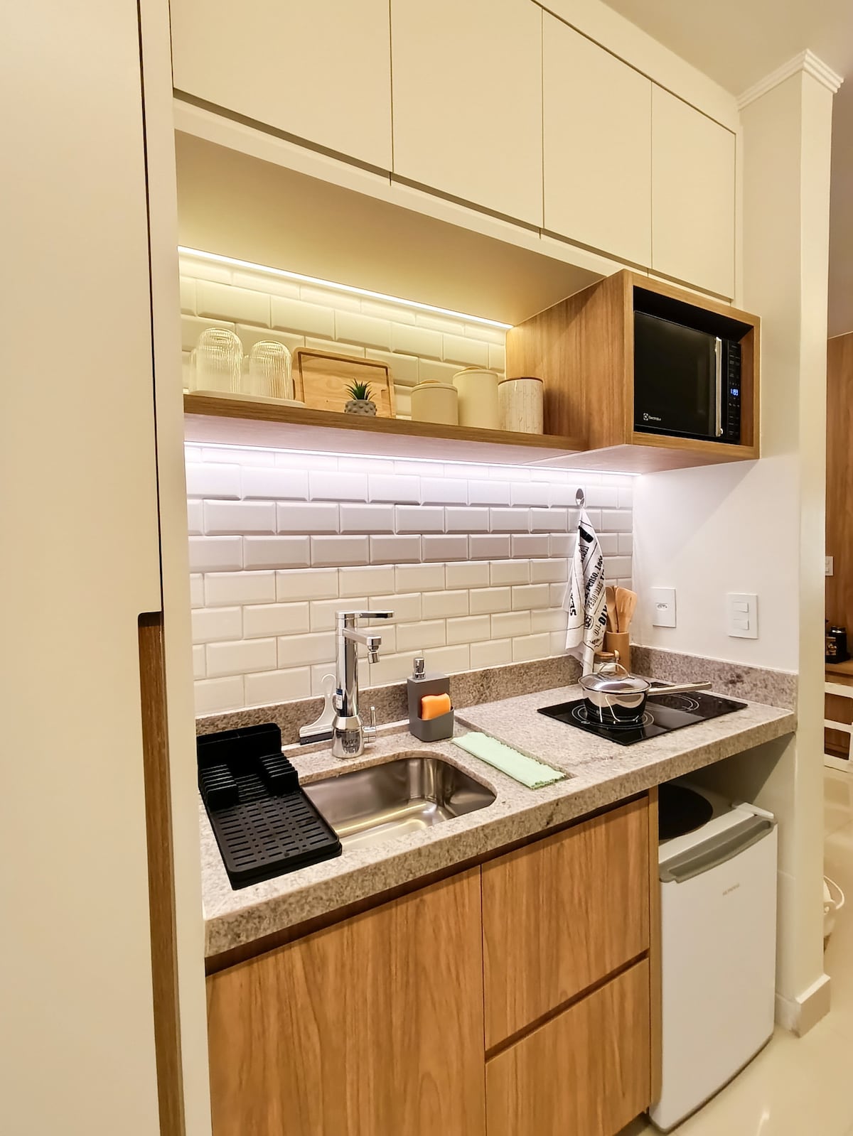 A compact kitchen area features light wood cabinetry with modern finishes. A stainless steel sink is complemented by a microwave and a small refrigerator. White subway tiles and under-cabinet lighting create a bright environment, while a cooktop, utensils, and a cutting board are neatly arranged.