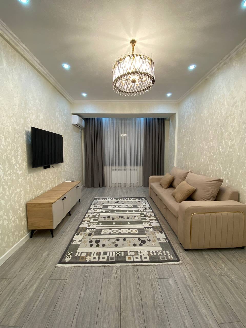 A well-lit living area features neutral tones with light-colored walls and sleek flooring. A contemporary sofa sits along one side, and a flat-screen television is mounted on the opposite wall. A decorative rug adds texture to the space, while drapes gently filter natural light.