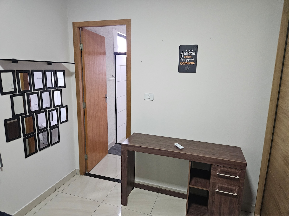 A modest workspace is shown with a wooden desk featuring a small drawer. A wall with a collage of framed pictures is visible, along with a doorway leading to another space. Natural light illuminates the area, enhancing its simplicity.