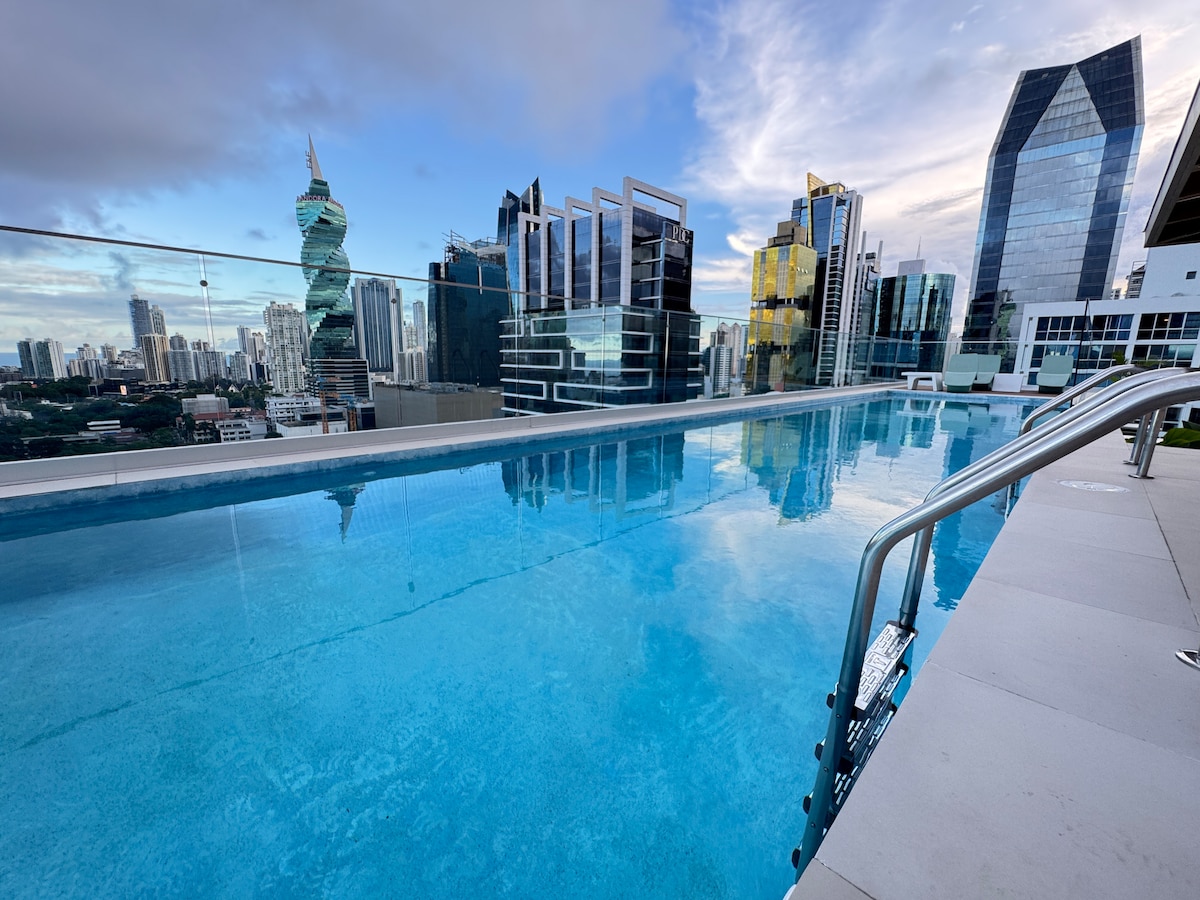 An infinity pool stretches along the rooftop, reflecting the skyline of Panama City. The clear blue water creates a seamless view of tall buildings and skyscrapers, with sunlight illuminating the area. Comfortable lounge chairs are positioned nearby, offering spaces for relaxation.