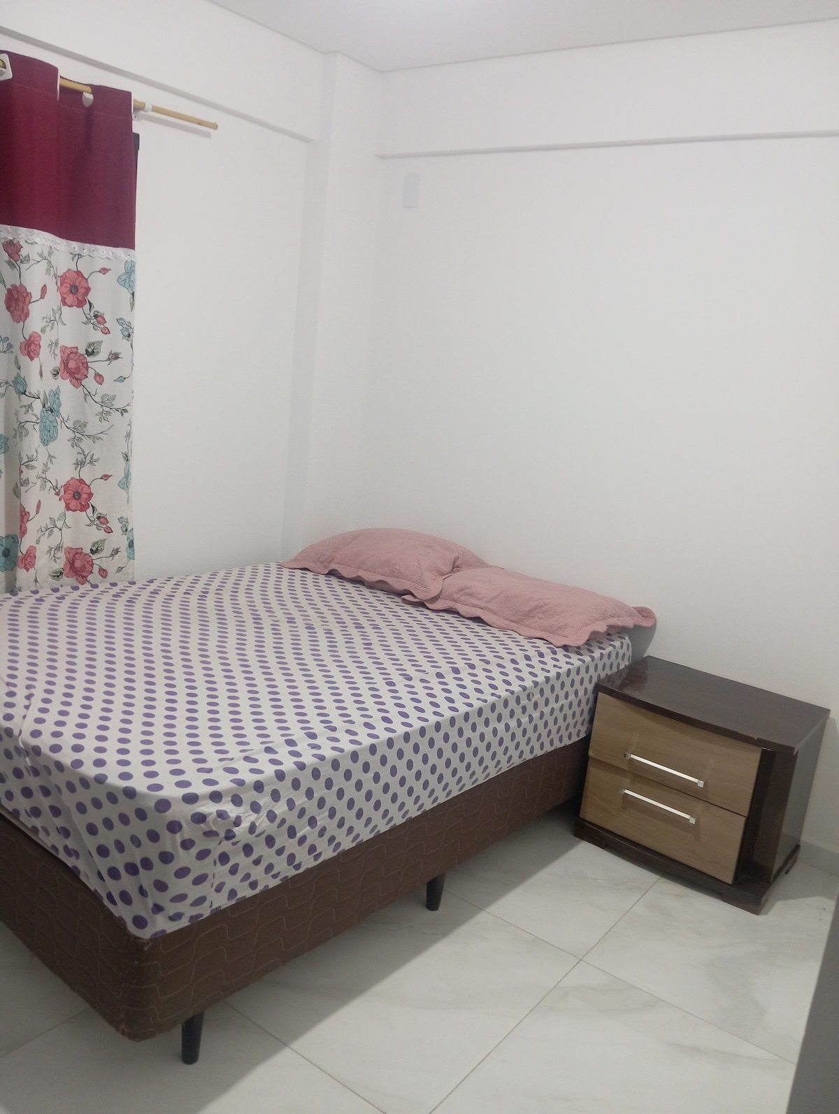A compact sleeping area features a bed with a polka-dot patterned mattress and soft bedding. A wooden nightstand stands beside the bed, equipped with two drawers. Natural light is filtered through floral curtains beside the bed, contributing to a calming environment.
