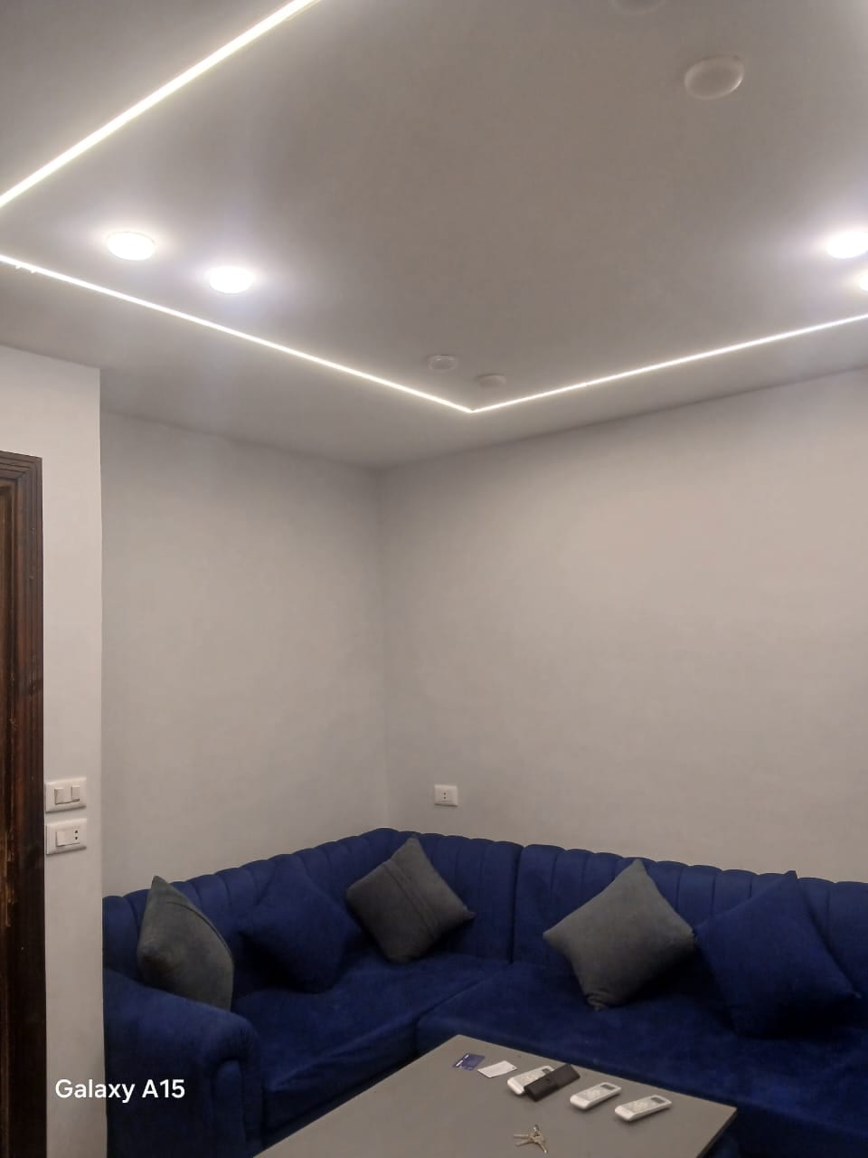 A seating area is featured with a blue, plush sofa adorned with several cushions. A light-colored coffee table is positioned in front, and the space is illuminated by modern recessed lighting along the ceiling. The walls are painted in a neutral hue.