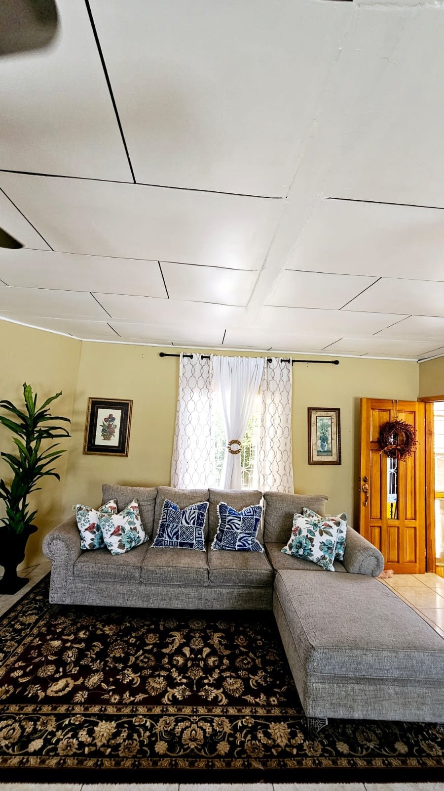 A cozy living area features a spacious gray sectional sofa adorned with patterned cushions. Natural light filters through a sheer curtain, illuminating decorative wall art and a potted plant. A wooden door with a wreath leads to the outside, adding to the welcoming feel of the space.