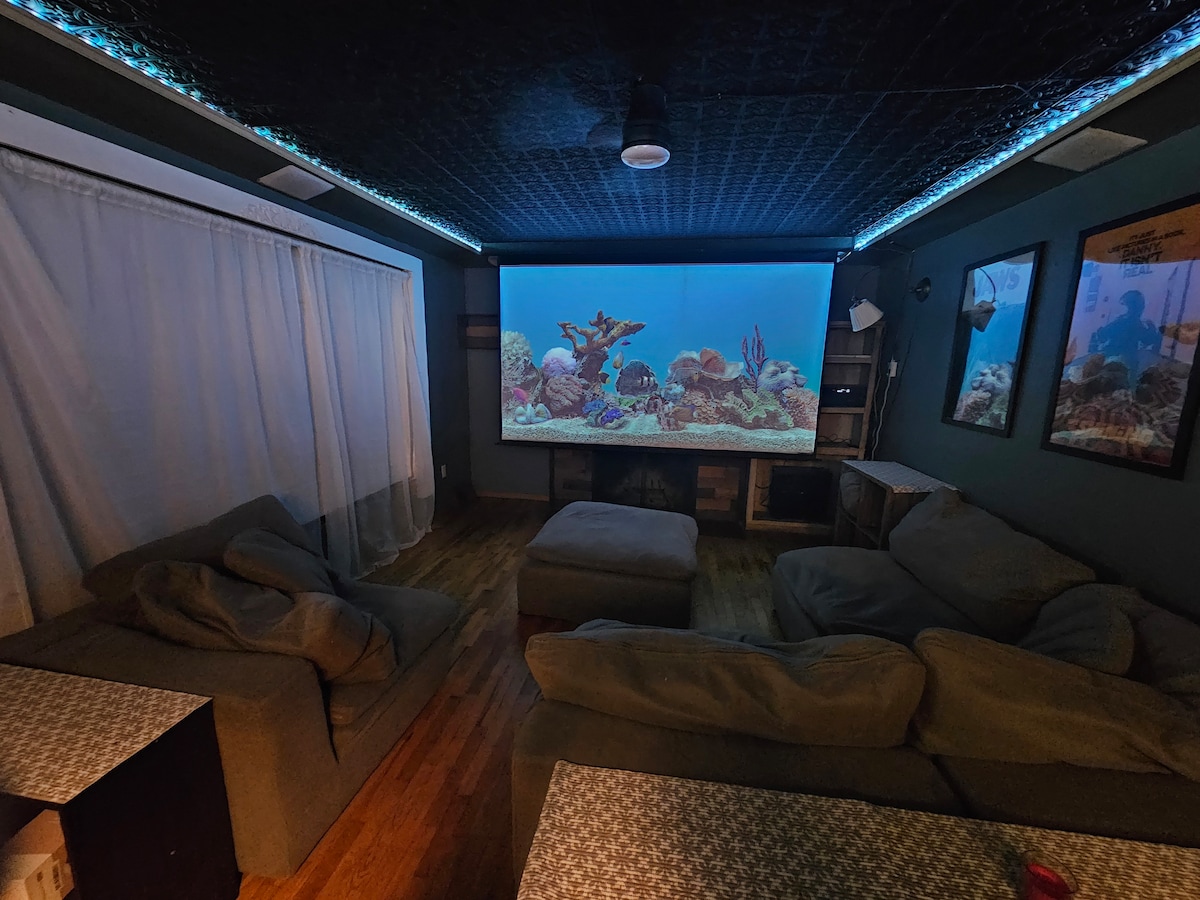 A home theater is equipped with a large projection screen displaying an underwater scene. Comfortable seating is arranged around a central ottoman, and soft lighting is provided by LED strips along the ceiling. Decorative wall art complements the room's cozy atmosphere.