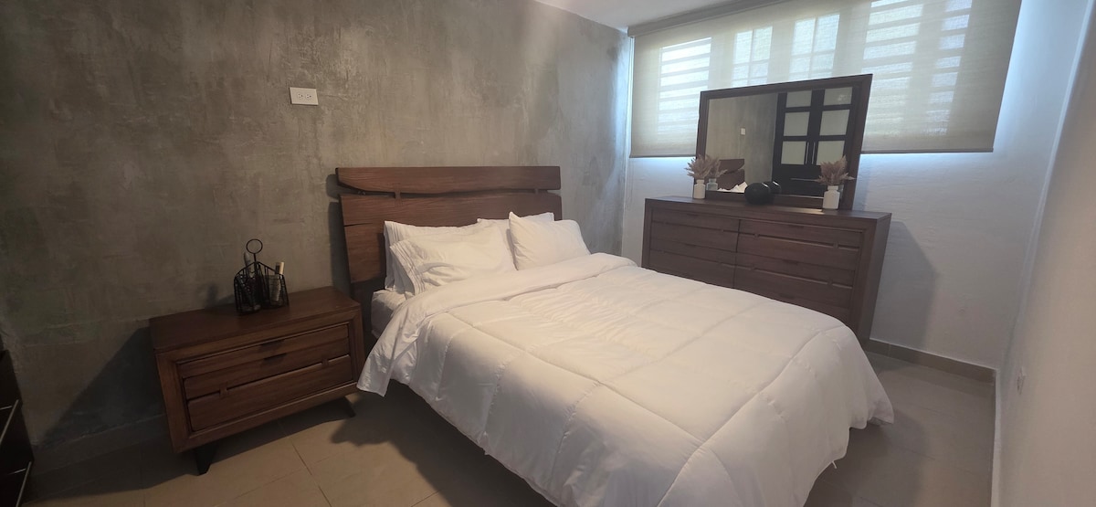 A modern bedroom features a queen bed with white linens, flanked by bedside tables. A dresser with a mirror stands opposite, and natural light filters through partially covered windows, highlighting a minimalist decor.