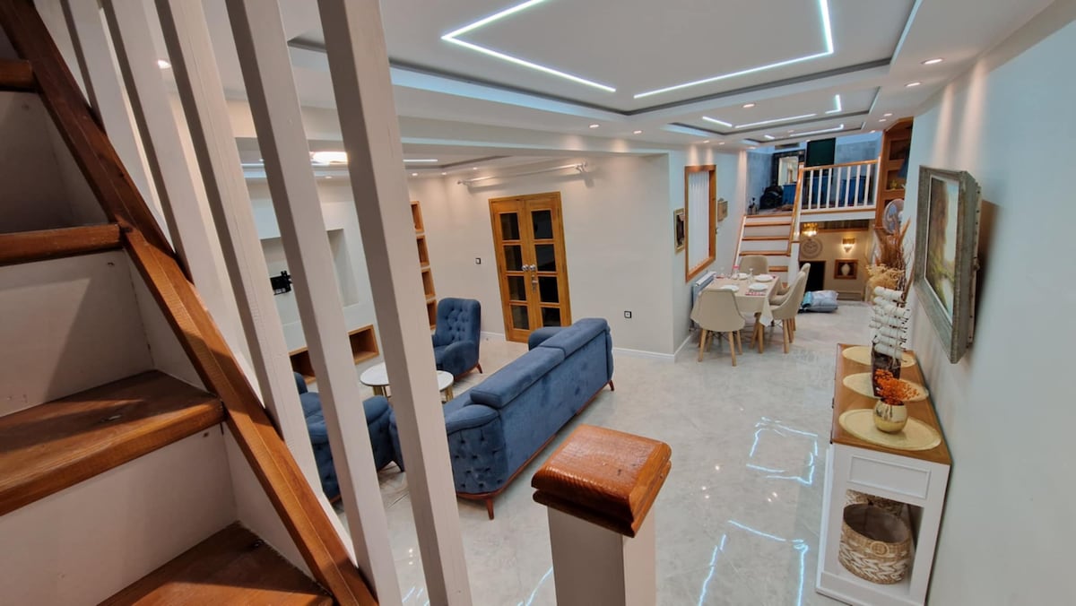 The interior space features an open layout with modern furnishings. A staircase is visible to the left, leading to the upper floor. A seating area with blue armchairs and a small table is positioned beside a dining area, creating an inviting and spacious atmosphere.