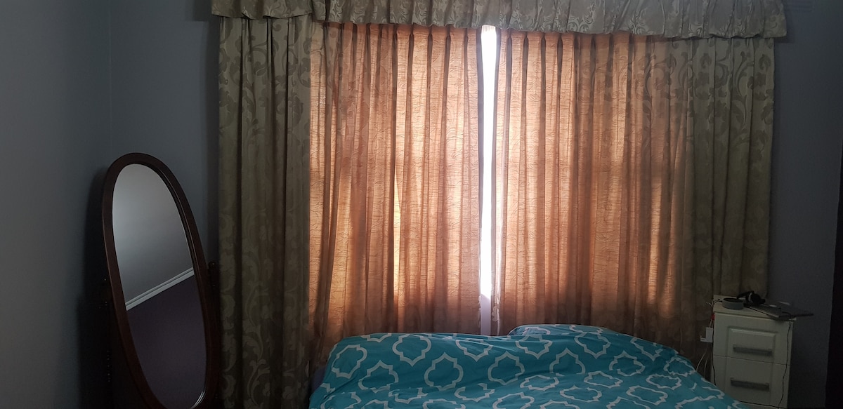 A cozy sleeping area features a spacious bed covered with a blue patterned blanket. Sunlight filters through the partially closed curtains, illuminating the room. A floor-length mirror and a nightstand are positioned nearby, contributing to a functional and inviting layout.