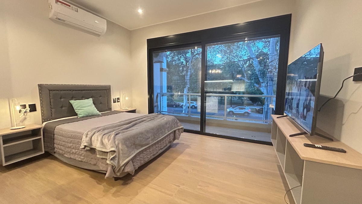 A spacious bedroom features a large bed with a grey upholstered headboard, complemented by a light blanket. Two bedside tables with lamps are positioned on either side. A television is mounted on the wall opposite the bed, and large sliding glass doors offer a view of the outside.