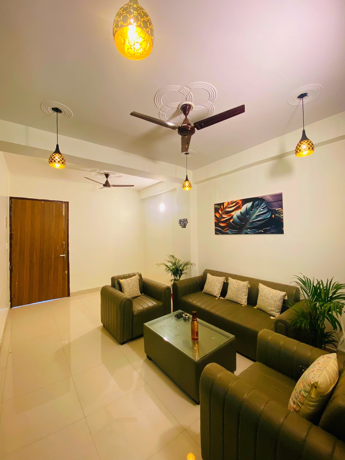 A modern living area features two green sofas arranged around a glass coffee table. Soft lighting is provided by hanging fixtures, while two ceiling fans circulate air. A piece of wall art adds a touch of color, and potted plants bring a hint of nature indoors.