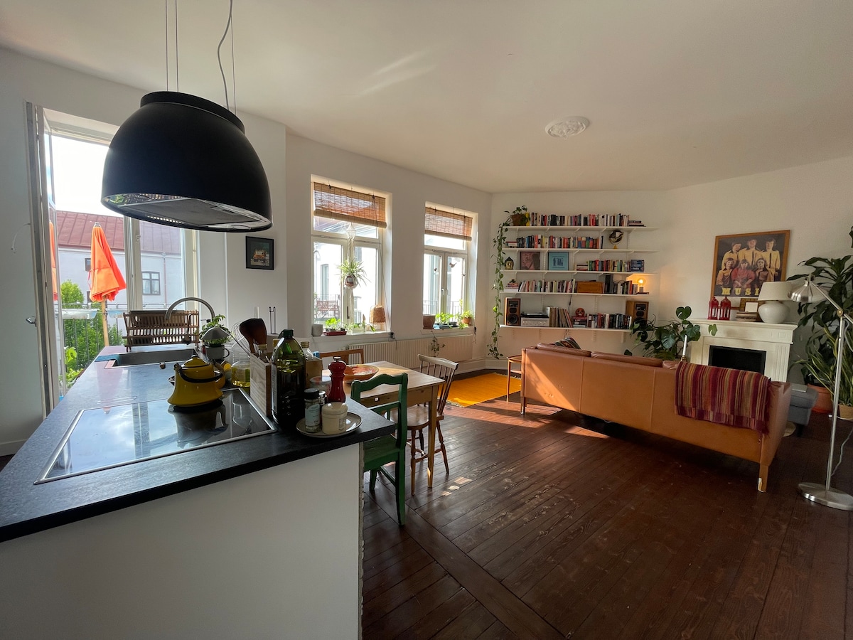 A bright living space with an open kitchen features modern appliances and a dining area with wooden chairs. A comfortable sofa is arranged against a wall lined with bookshelves, while large windows allow natural light to fill the room. Indoor plants add a touch of greenery.