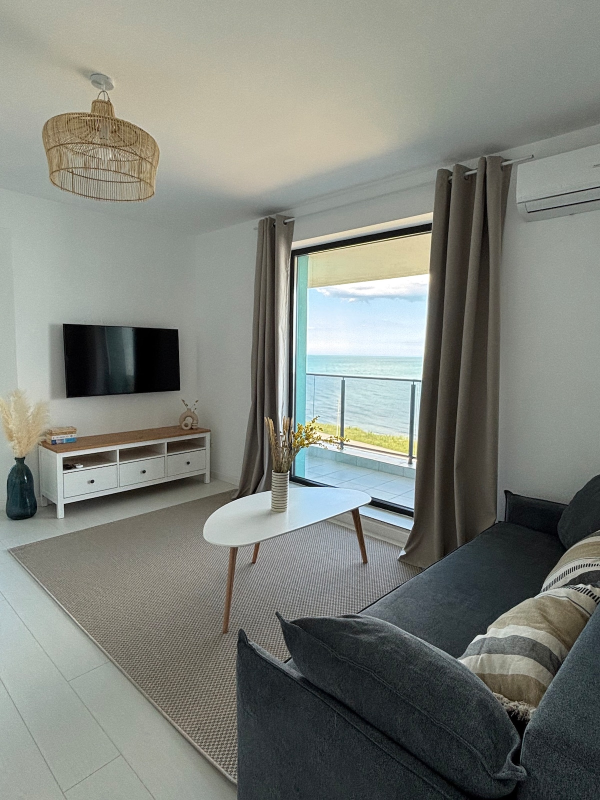 A bright living area features a spacious gray sofa and a light wood coffee table, positioned on a textured area rug. Large windows provide a sea view and natural light, while air conditioning is visible above the sleek media console housing a television.