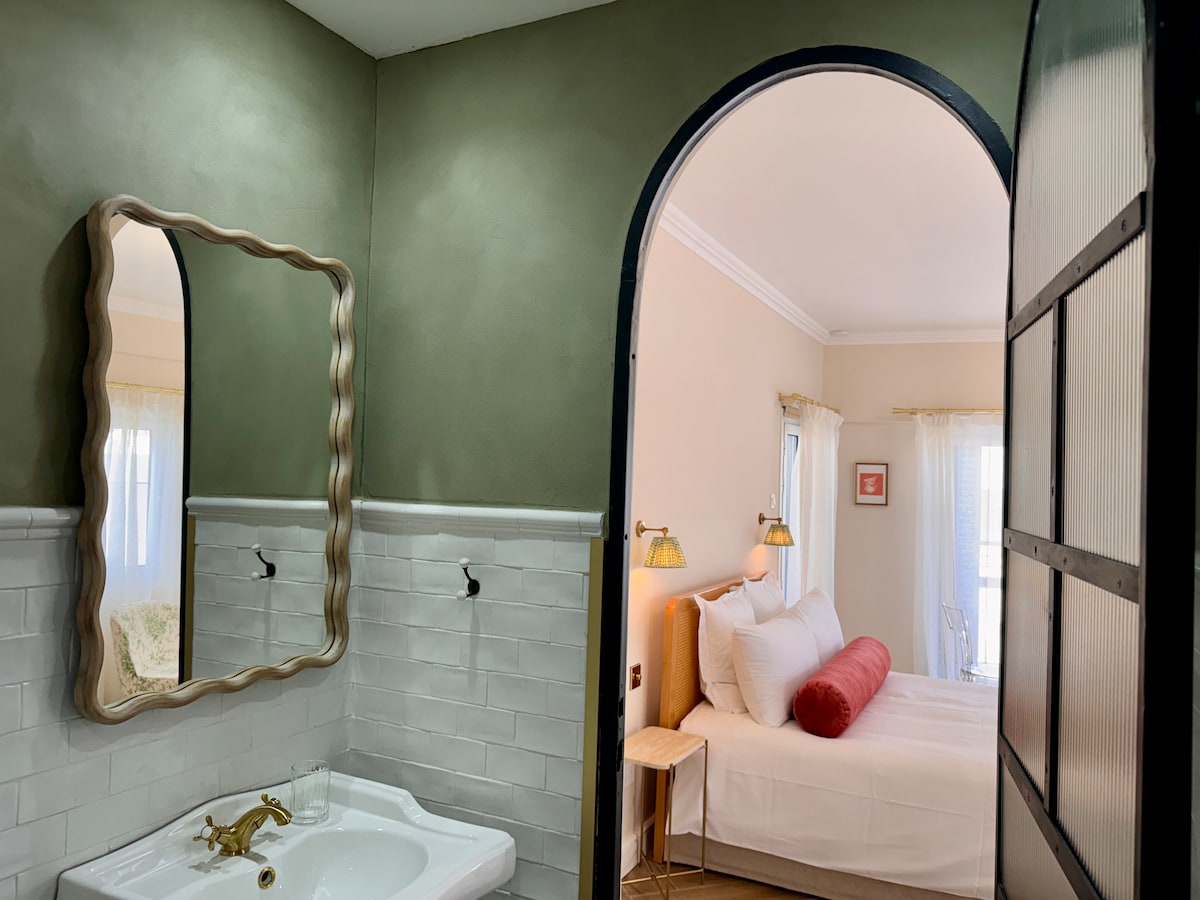 A view into a room is offered through an arched doorway. The light-colored room is illuminated by natural light. A spacious bed with white linens is seen, complemented by a decorative red pillow. A stylish mirror hangs on the wall with a unique frame.