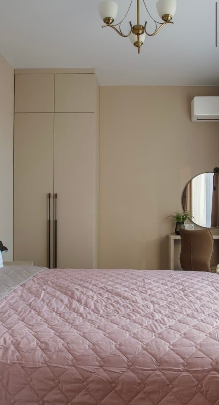 A cozy bedroom features a spacious bed covered with a soft quilt in subtle pink tones. A modern round mirror and a chair are placed near a window, allowing natural light to illuminate the warm walls. A sleek wardrobe stands against one wall.