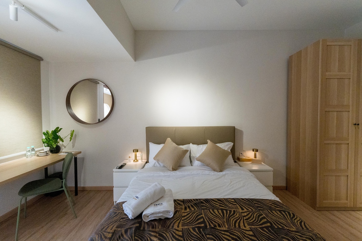 A well-appointed sleeping area features a spacious bed with soft linens and decorative pillows. A work desk with a plant is positioned nearby, alongside warm lighting and a round mirror. Soft neutral tones and wooden accents create a calming ambiance.