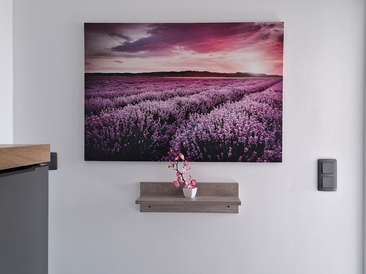 A decorative wall art piece is displayed above a wooden shelf, depicting a vibrant lavender field under a soft pink sky. A small potted plant is placed on the shelf, adding a touch of nature to the space.