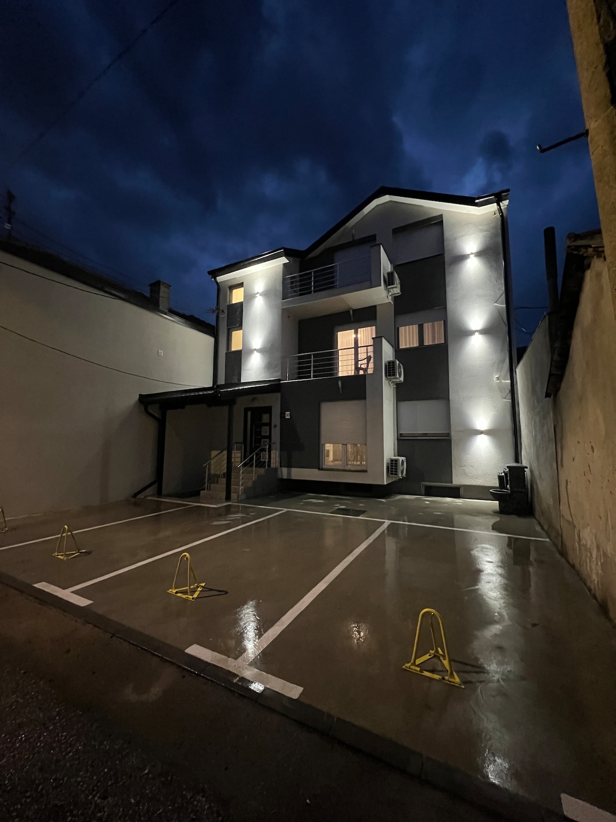 The building's exterior is illuminated by bright lights, highlighting the modern design and multiple levels. An entrance area is visible, with a covered porch leading to the front door. A small parking area is marked with visible parking spaces, reflecting light in the evening environment.