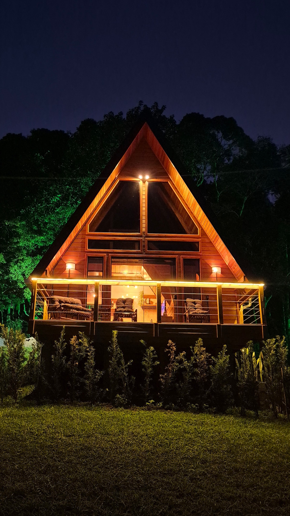 A triangular-shaped cabin is illuminated from within, showcasing a welcoming interior. Surrounding greenery is highlighted in soft light, creating a serene environment. The structure features large windows that enhance the connection with nature and a cozy outdoor area with well-maintained shrubs.