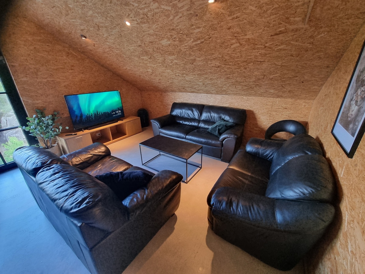 A cozy seating area features two leather sofas facing a coffee table, providing ample space for relaxation. A wall-mounted smart TV is visible, with natural light filtering in through large windows. The walls showcase a textured finish that adds warmth to the space.