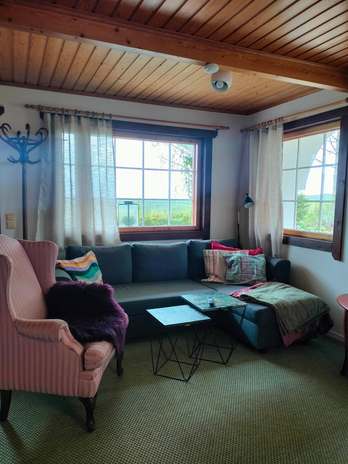 A cozy living room area features a dark sectional sofa complemented by colorful cushions and a soft throw. Natural light enters through large windows adorned with sheer curtains. A small glass table rests between the seating, supported by thin metal legs, enhancing the inviting space.