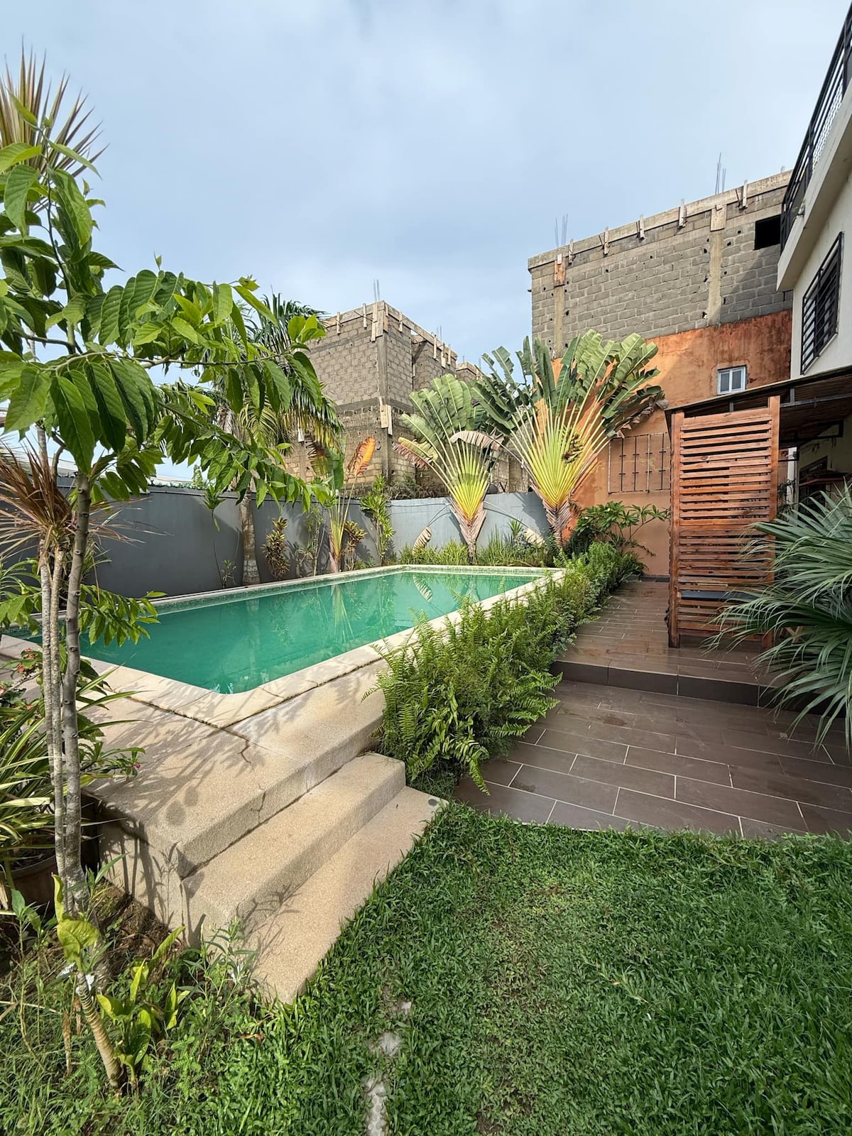 A tranquil pool is surrounded by lush greenery, featuring well-maintained landscaping with tropical plants. The poolside area includes steps leading into the water, and a wooden privacy screen enhances the serene outdoor setting.