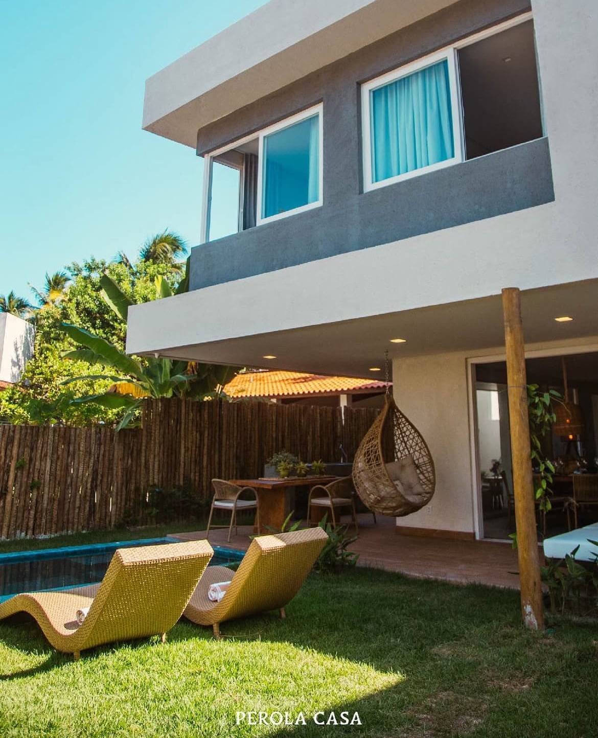 A two-story house is depicted, featuring large windows and a shaded area adjacent to lush greenery. A pool is visible in the foreground, with two lounge chairs positioned on the grass. A hanging chair is seen in a covered patio space adjoining a dining area.