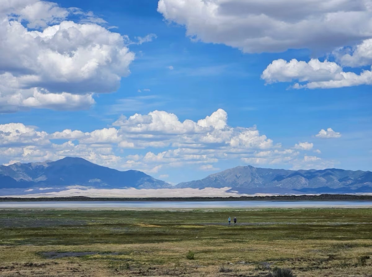A wide landscape features a vibrant green foreground, gently rolling hills, and distant mountains. Fluffy white clouds occupy a bright blue sky, creating a serene backdrop. A strip of white sand can be seen along the base of the mountains, adding contrast to the scenic view.