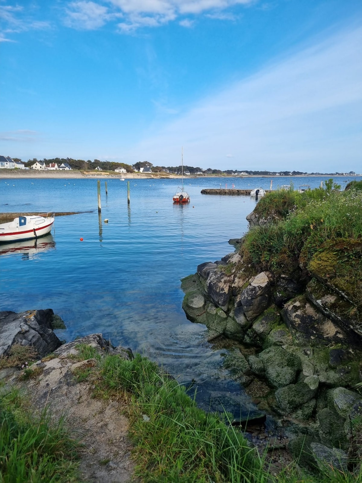 A serene coastal scene is depicted, featuring calm waters reflecting a clear blue sky. Small boats are anchored nearby, while rocky shores and patches of greenery frame the view. The distant shoreline shows hints of residential buildings amidst the natural landscape.